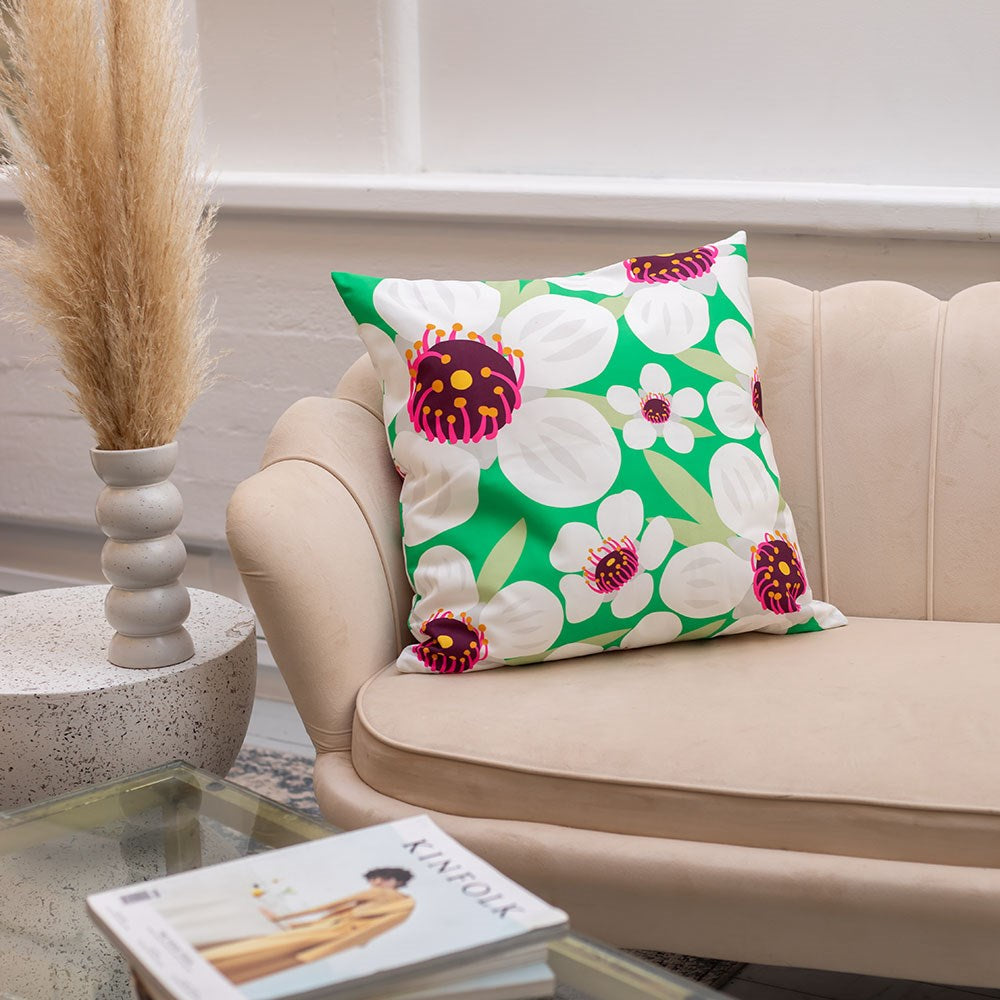 A beige couch displays the Bright Botanical Manuka Cushion Cover, featuring a green, white, and purple floral pattern. Made of polyester for indoor or outdoor decor. A decorative vase with pampas grass sits on a nearby round table.
