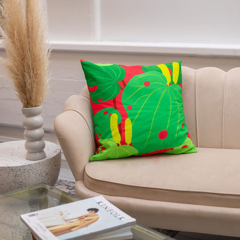 The Bright Botanical Kawakawa Cushion Cover, featuring a green and red tropical leaf print, rests on a beige sofa next to dried pampas grass. Ideal for indoor or outdoor use, it adds stylish flair beside a glass coffee table and magazine.