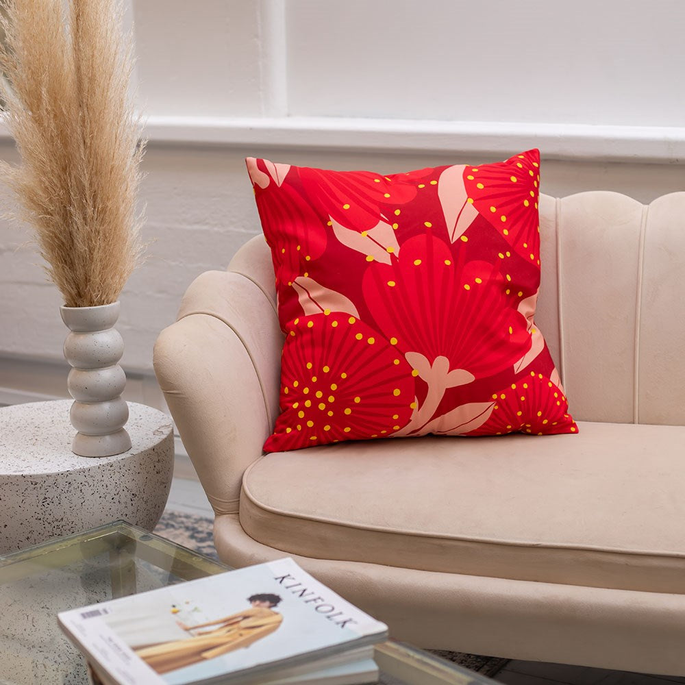 A beige couch is accented with the Bright Botanical Red Pohutukawa Cushion Cover. Nearby, a round table holds a pampas grass vase, and an open Kinfolk magazine sits atop the glass coffee table.