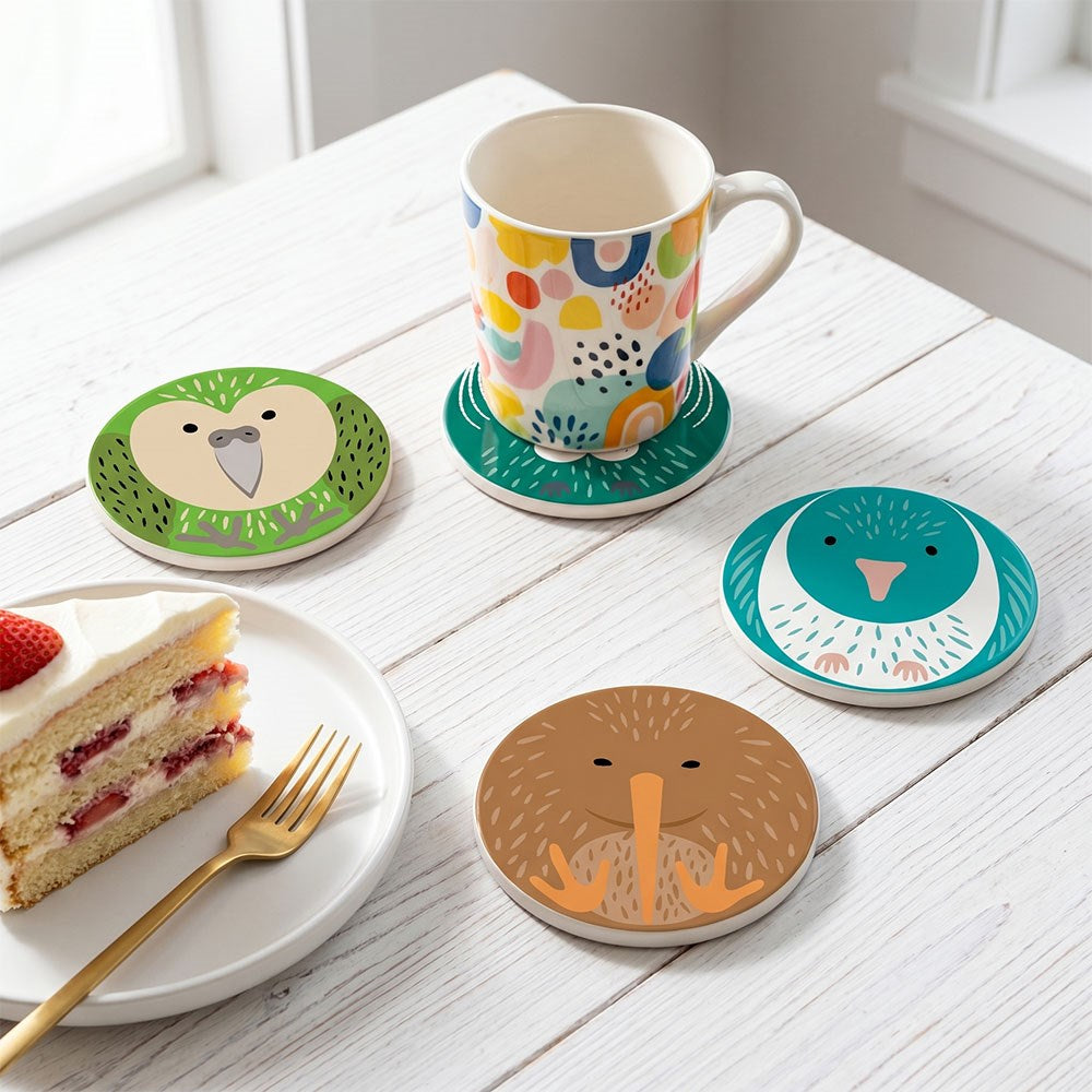 A white table with a slice of layered cake on a plate with a gold fork, a patterned mug on a colorful coaster, and three more Set of 4 Cuties Ceramic Coasters featuring illustrated New Zealand birds.