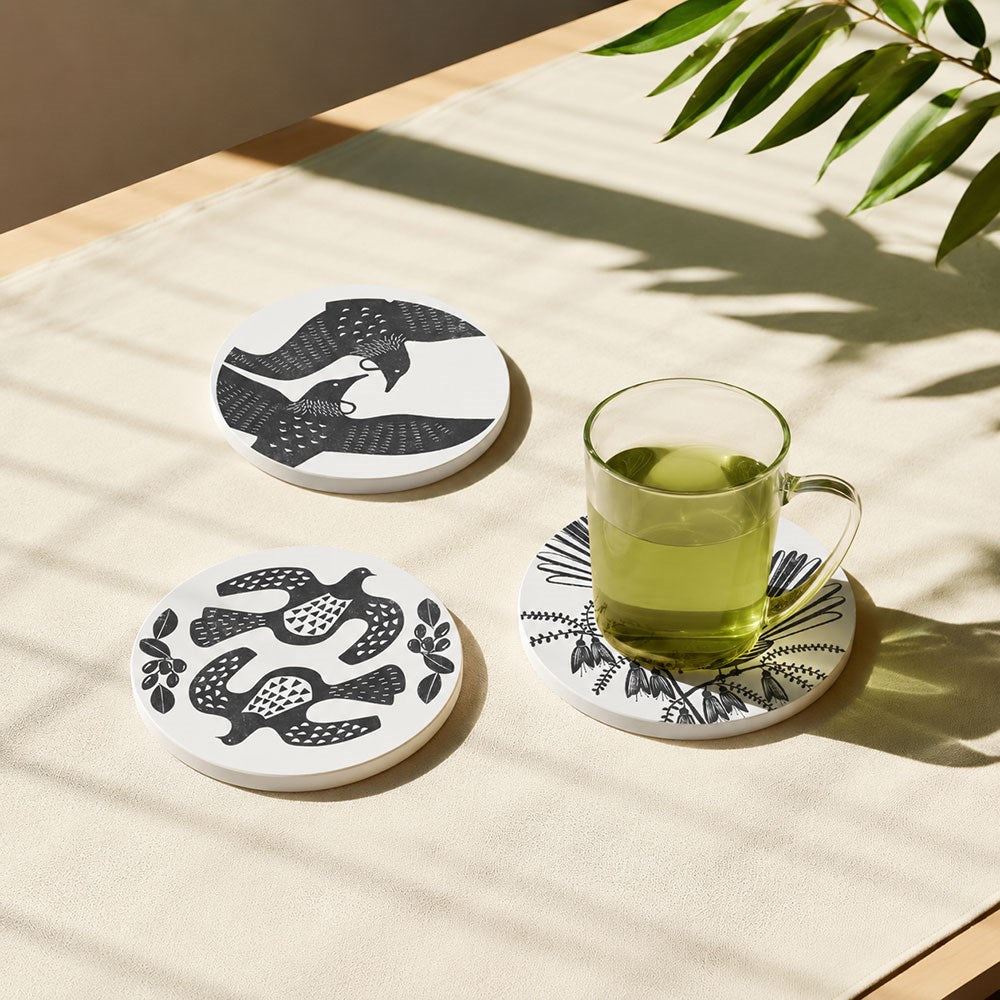 A glass mug with green liquid rests on a Set of 3 Linocut Ceramic Coasters—featuring black-and-white patterns and New Zealand bird designs—on a light tablecloth beneath natural light and leafy shadows.