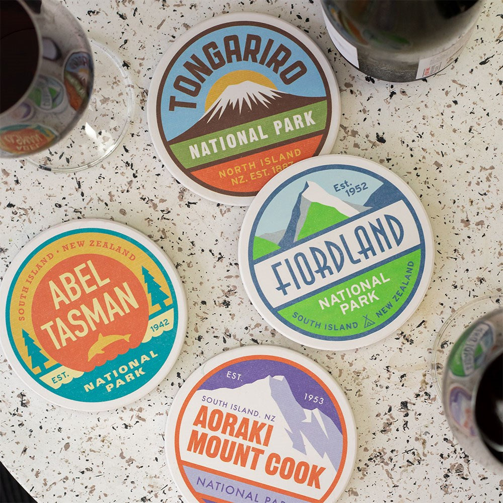 Set of 4 National Park Coasters by Glenn Jones, featuring colorful designs of Tongariro, Fiordland, Abel Tasman, and Aoraki Mount Cook, displayed on a speckled surface near glasses of red wine.
