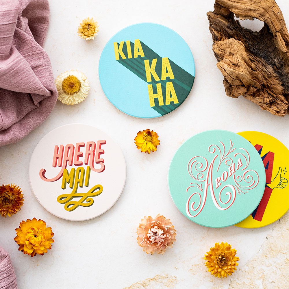 The Set of 4 Te Reo Ceramic Coasters features bold pop art designs and phrases like KIA KAHA, HAERE MAI, and Aroha. Displayed on a white surface with dried flowers, wood, and a mauve cloth.