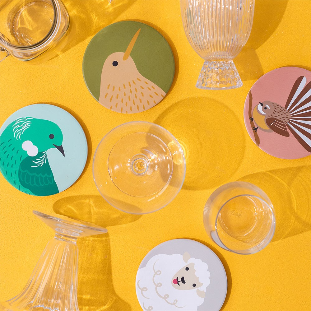 The Set of 4 Storybook Ceramic Coasters, each printed in New Zealand and featuring a bird, sheep, pigeon, and pheasant design, is shown on a yellow surface with assorted empty glass cups around them.