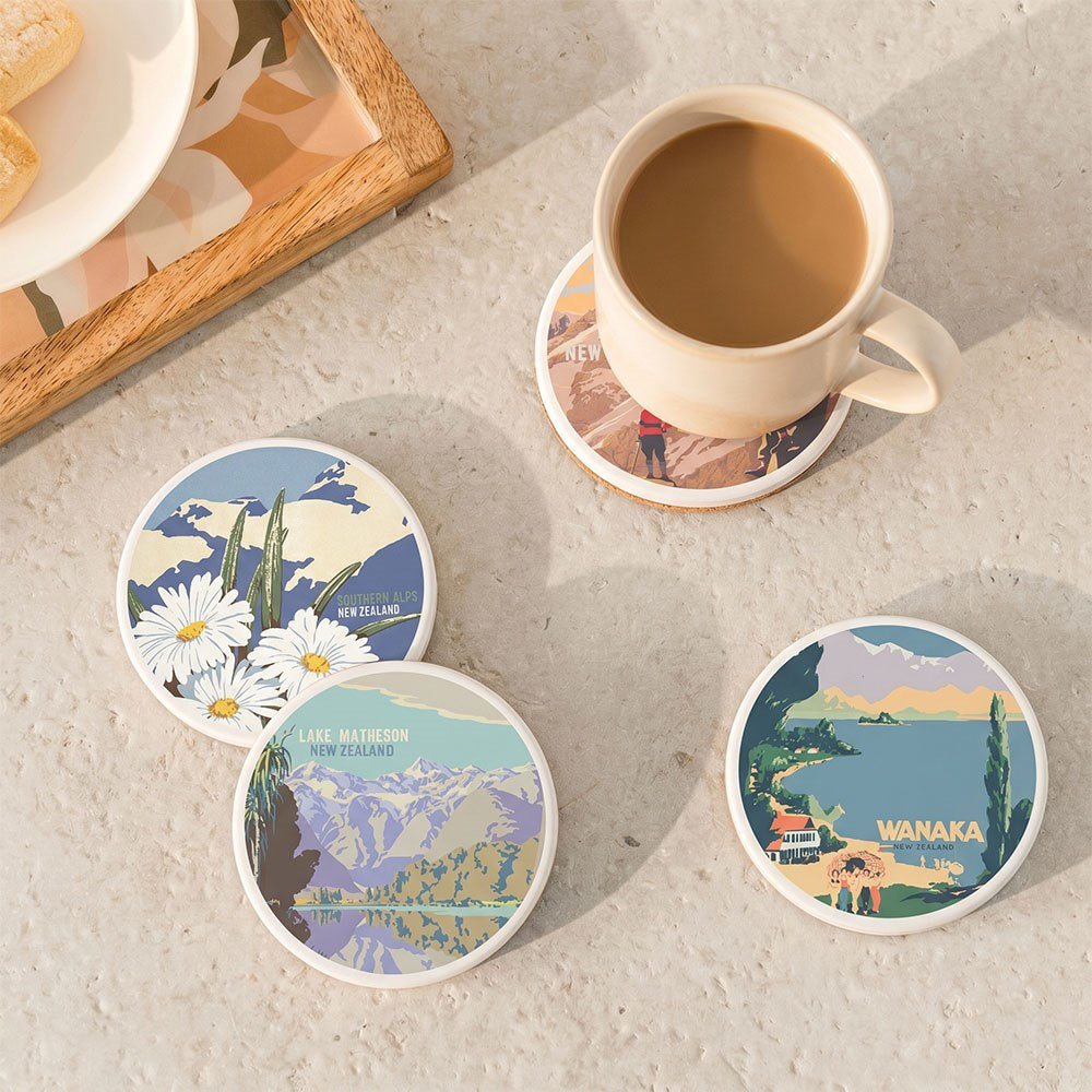 A beige mug of coffee rests on a coaster from the Set of 4 Tourist Ceramic Coasters, which features scenic, vintage-inspired New Zealand illustrations. Three more coasters from the set are nearby on a light, textured surface.