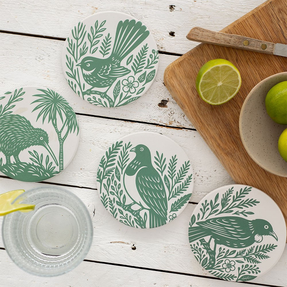 A Set of 4 Woodcut Ceramic Coasters with green bird illustrations is displayed on a white wooden table with a glass of lemon water, a cutting board, knife, and fresh limes. Coasters are printed in New Zealand.
