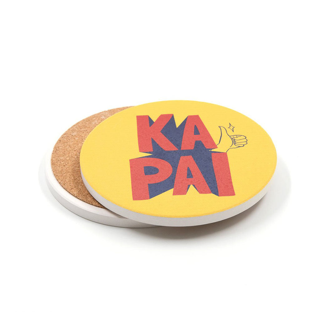 This Set of 4 Te Reo Ceramic Coasters features round, pop art-inspired designs with a yellow top, bold red and purple "KA PAI" typography, a thumbs-up graphic, and a brown cork back—perfect for New Zealand-themed decor.