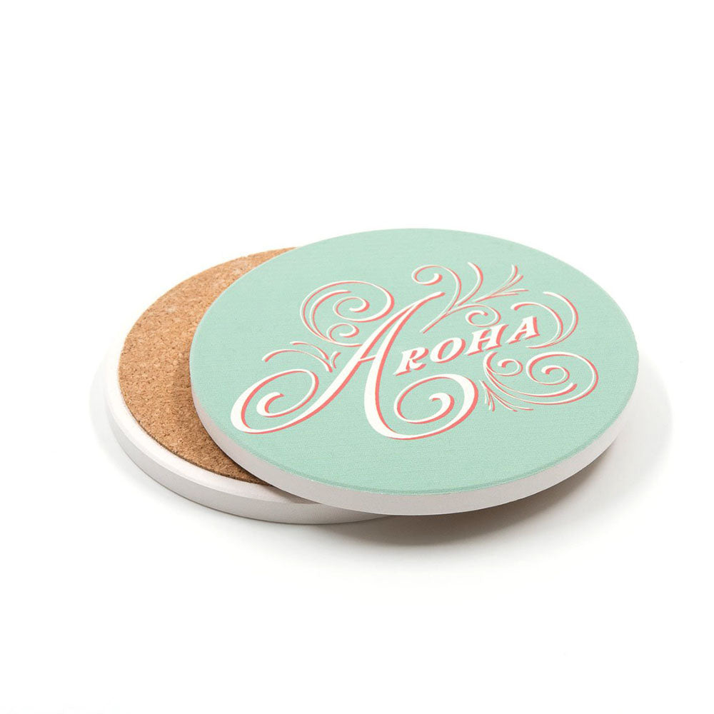 A set of 4 Te Reo ceramic coasters with cork bases and light green tops, each featuring "Aloha" in decorative script inspired by pop art. Coasters are shown partially stacked for a stylish display.
