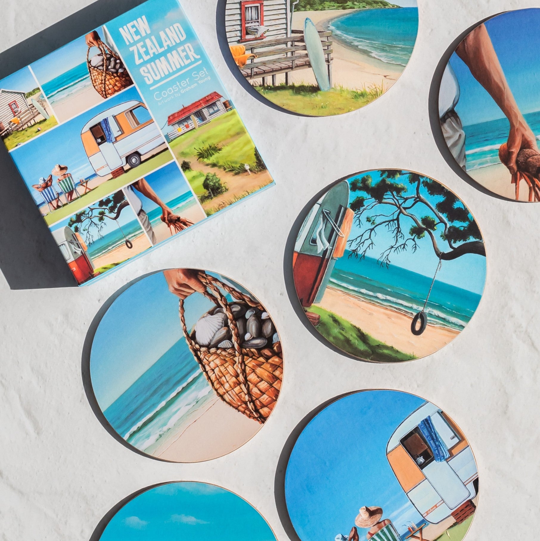 The Graham Young NZ Summer Set of 6 Coasters, each with beach, caravan, and woven basket illustrations, is arranged on a white surface beside its labeled box. Ideal for beach-lovers seeking vibrant summer-themed decor.