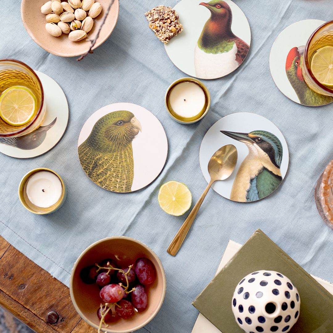 A table set with a Hushed Birds Set of 6 Coasters featuring New Zealand native birds, a bowl of grapes, pistachios, lit candles, lemon water with a slice, a gold spoon, and a polka-dot container on a blue cloth.