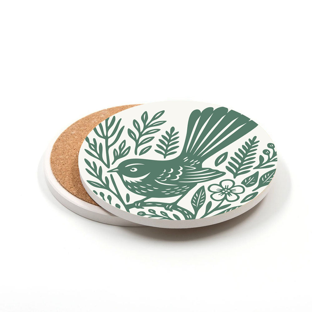 A round ceramic coaster from the Set of 4 Woodcut Ceramic Coasters features a green woodcut-style bird with leaves and flowers on white, cork-backed, with another coaster partially visible underneath.