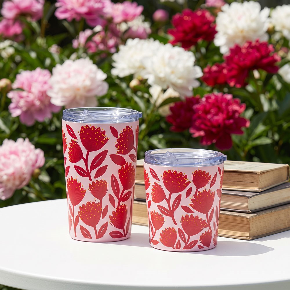Two lidded Pohutukawa Glade Coffee Cups with vacuum insulation and red floral designs sit on a white table next to stacked books. Pink and white flowers, along with greenery, appear slightly out of focus in the background.