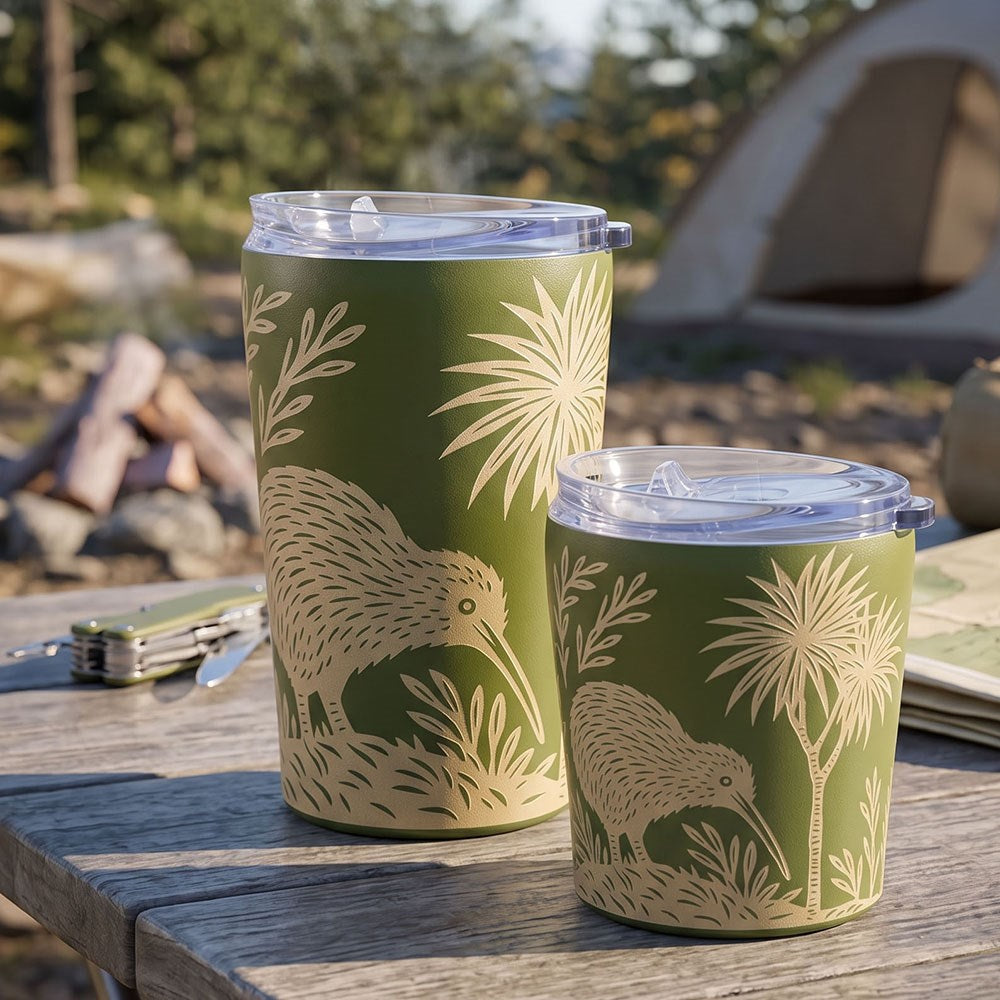 Two green Woodcut Kiwi Coffee Cups with cream-colored kiwi bird and plant designs sit on a wooden outdoor table. A tent, campfire, and trees blur in the background, evoking a camping atmosphere. These stainless steel, vacuum-insulated travel mugs are perfect for keeping your drinks hot or cold while you enjoy the outdoors.