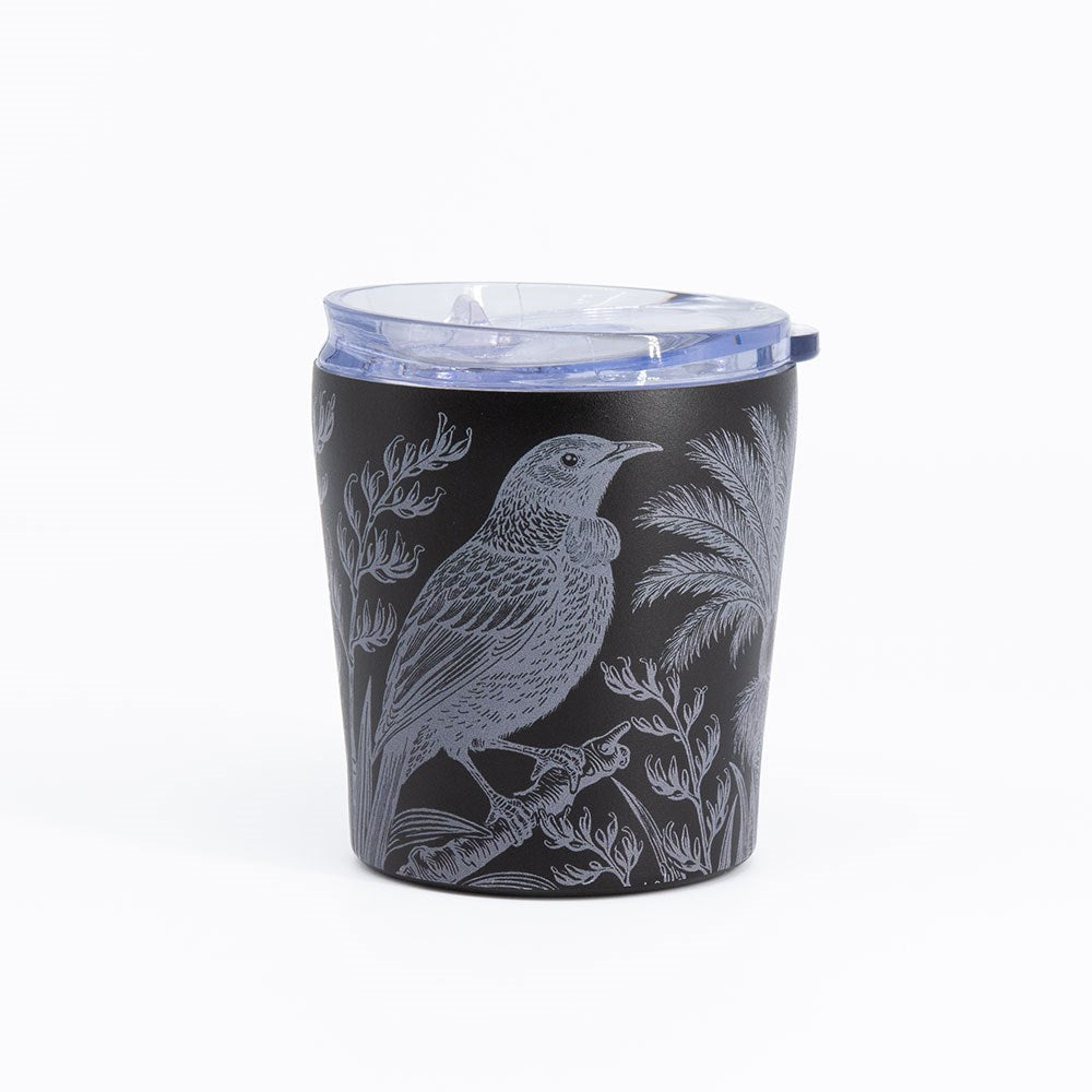 Introducing the Midnight Tui Coffee Cup—a black stainless steel travel mug with a clear plastic lid, adorned with a detailed illustration of a tui bird perched on a branch among foliage and palm trees in light gray. This vacuum-insulated coffee cup is spill resistant and set against a plain white background.