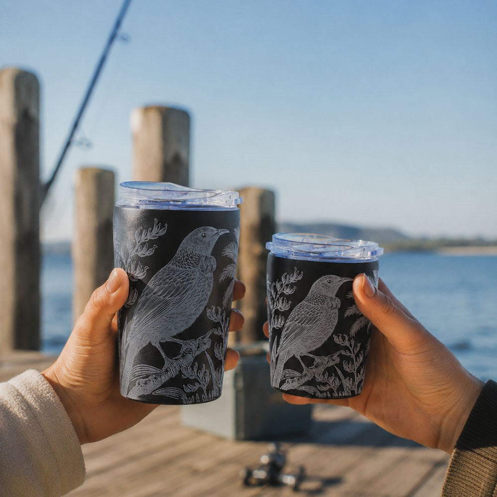 Two people hold stylish, spill-resistant Midnight Tui Coffee Cups, featuring white bird and plant designs on stainless steel. They are standing on a wooden dock near the water, with a fishing rod and reel visible in the background under a clear blue sky.