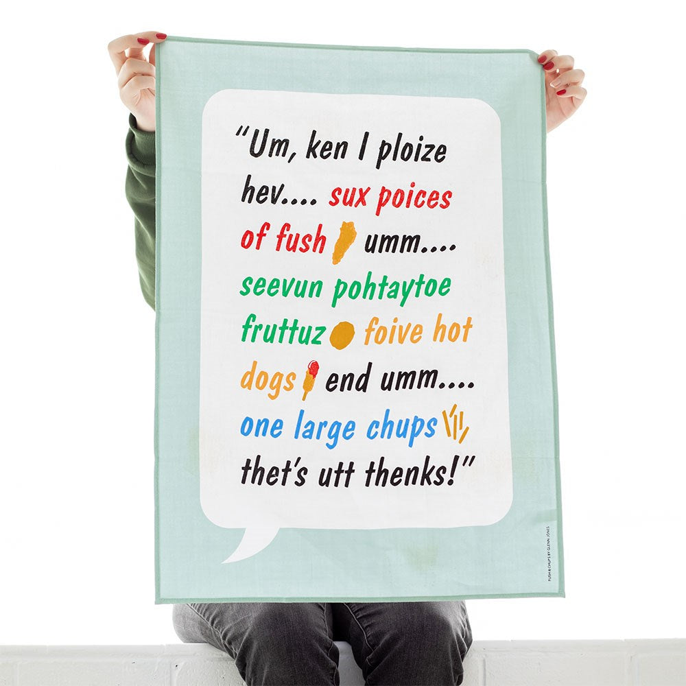 A person holds up the Glenn Jones - Fush & Chups Tea Towel, designed in New Zealand, displaying colorful fonts with a humorous phonetic artwork inspired by “Um, can I please have six pieces of fish…”.