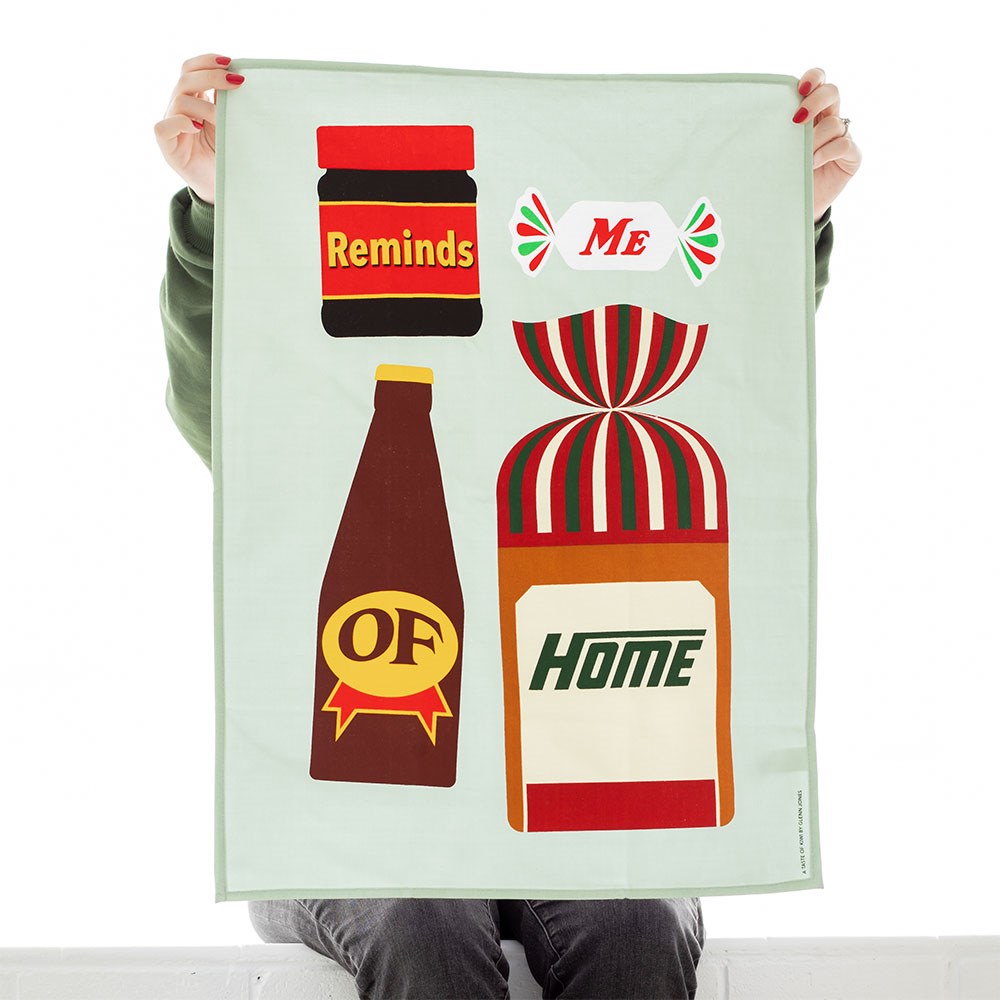 A person seated and holding up the Glenn Jones - Taste of Kiwi Tea Towel, which features graphic illustrations of a jar labeled Reminds, candy labeled Me, bottle labeled Of, and bread labeled Home. Designed in New Zealand by artist Glenn Jones.