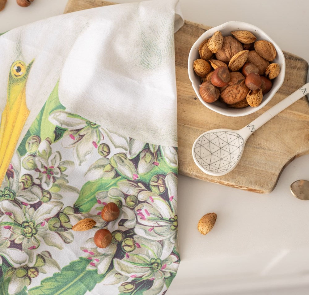 A wooden board with a bowl of mixed nuts and a spoon rests partly covered by the Birds & Botanicals Heron Tea Towel, as several nuts are scattered on the towel and white surface below.