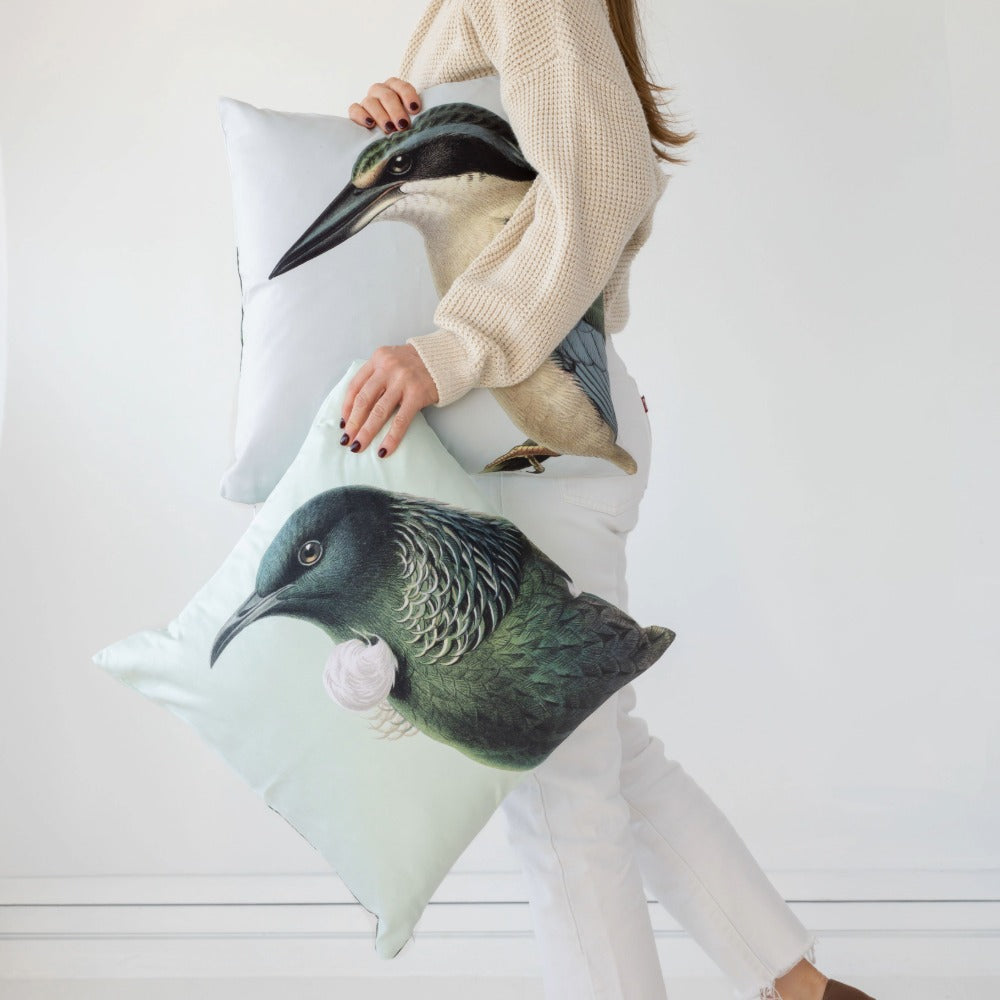 A person in a beige sweater and white pants holds two Hushed Green Tui Cushion Covers, each featuring a realistic illustration of New Zealand native birds, against a plain white background.