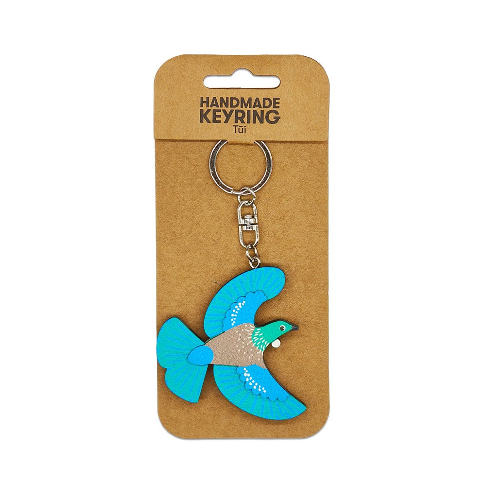 The Tui Wooden Keyring is a handmade wooden keychain shaped like a blue and brown tūī bird, attached to a metal ring. Packaged on brown card labeled HANDMADE KEYRING Tūī, its a unique Aotearoa keepsake.