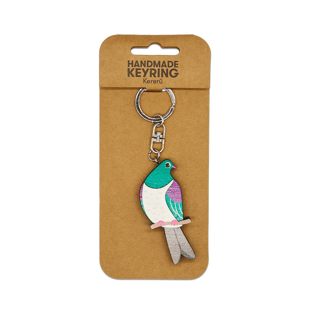 The Kereru Wooden Keyring features a handmade, colorful bird in green, white, pink, and purple on a metal keyring with chain, displayed on cardboard backing. This Kiwiana item is labeled Kereru.