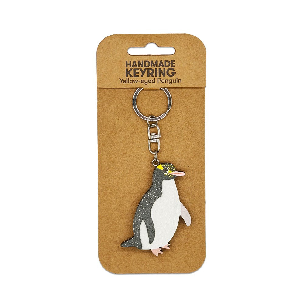 The Yellow-eyed Penguin Wooden Keyring is a handmade wooden keychain with a flat, cartoon-style yellow-eyed penguin design, attached to a metal ring and displayed on brown packaging labeled “Yellow-eyed Penguin Wooden Keyring.”.