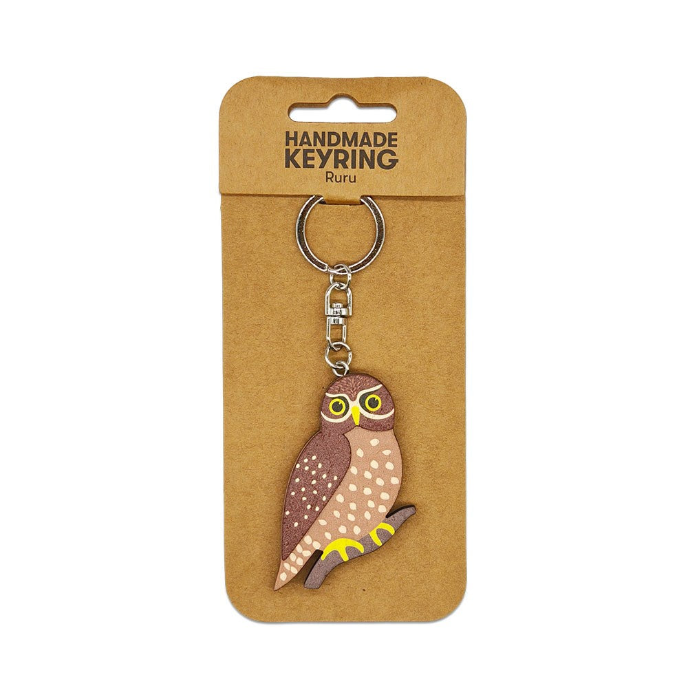 The Ruru Wooden Keyring is a handmade wooden keychain featuring a charming owl on a metal ring and chain, displayed on brown packaging labeled HANDMADE KEYRING Ruru—a unique Aotearoa keepsake.