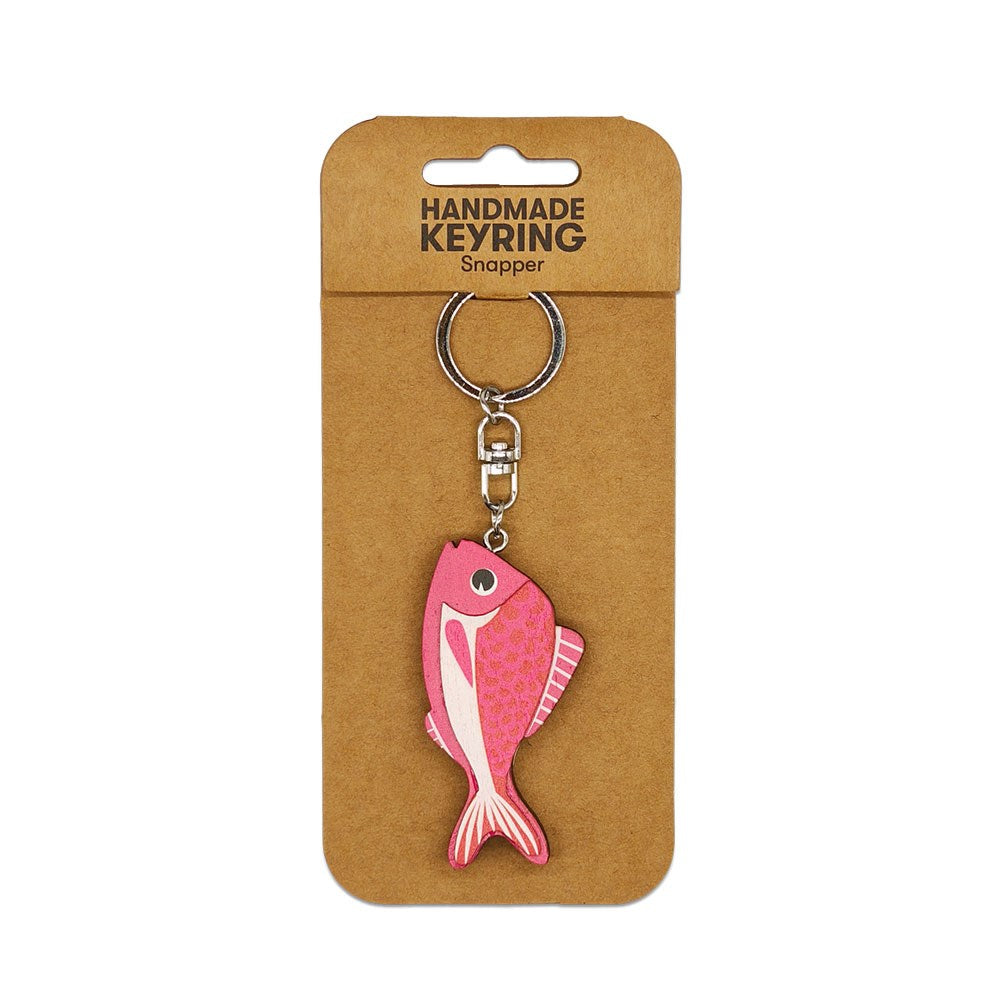 The Snapper Wooden Keyring is a pink, snapper-shaped wooden accessory with a metal clasp, attached to brown cardboard packaging labeled Handmade Keyring Snapper—a perfect handmade keychain for fish lovers.
