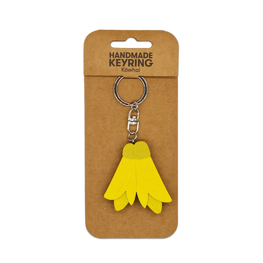 A Kowhai Wooden Keyring, shaped like a yellow flower and handmade from wood, is attached to a metal keychain and displayed on brown cardboard labeled HANDMADE KEYRING Kowhai.