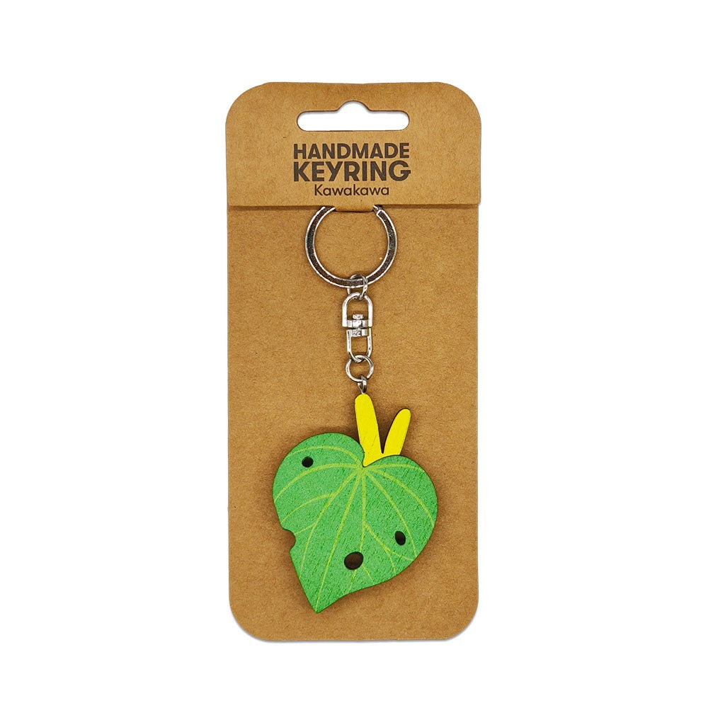 The Kawakawa Wooden Keyring is a handmade Kiwiana keychain featuring a green kawakawa design with black spots and a yellow stem on a silver keyring, displayed on brown cardboard labeled “Handmade Keyring Kawakawa.”.