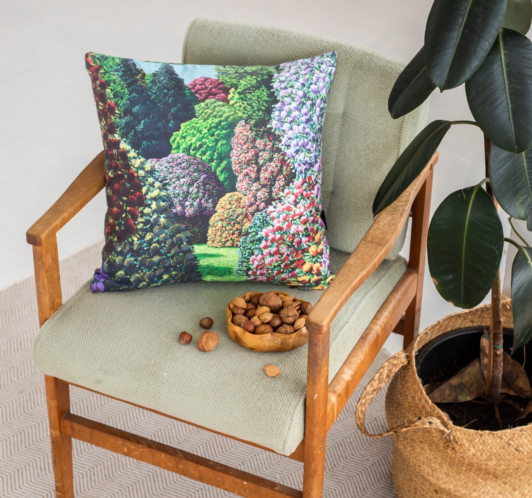A wooden armchair with a light green Karl Maughan - Forest Hill Cushion Cover holds a garden-inspired decorative pillow. A bowl of nuts and some scattered nuts are on the seat, while a potted plant in a woven basket sits nearby.