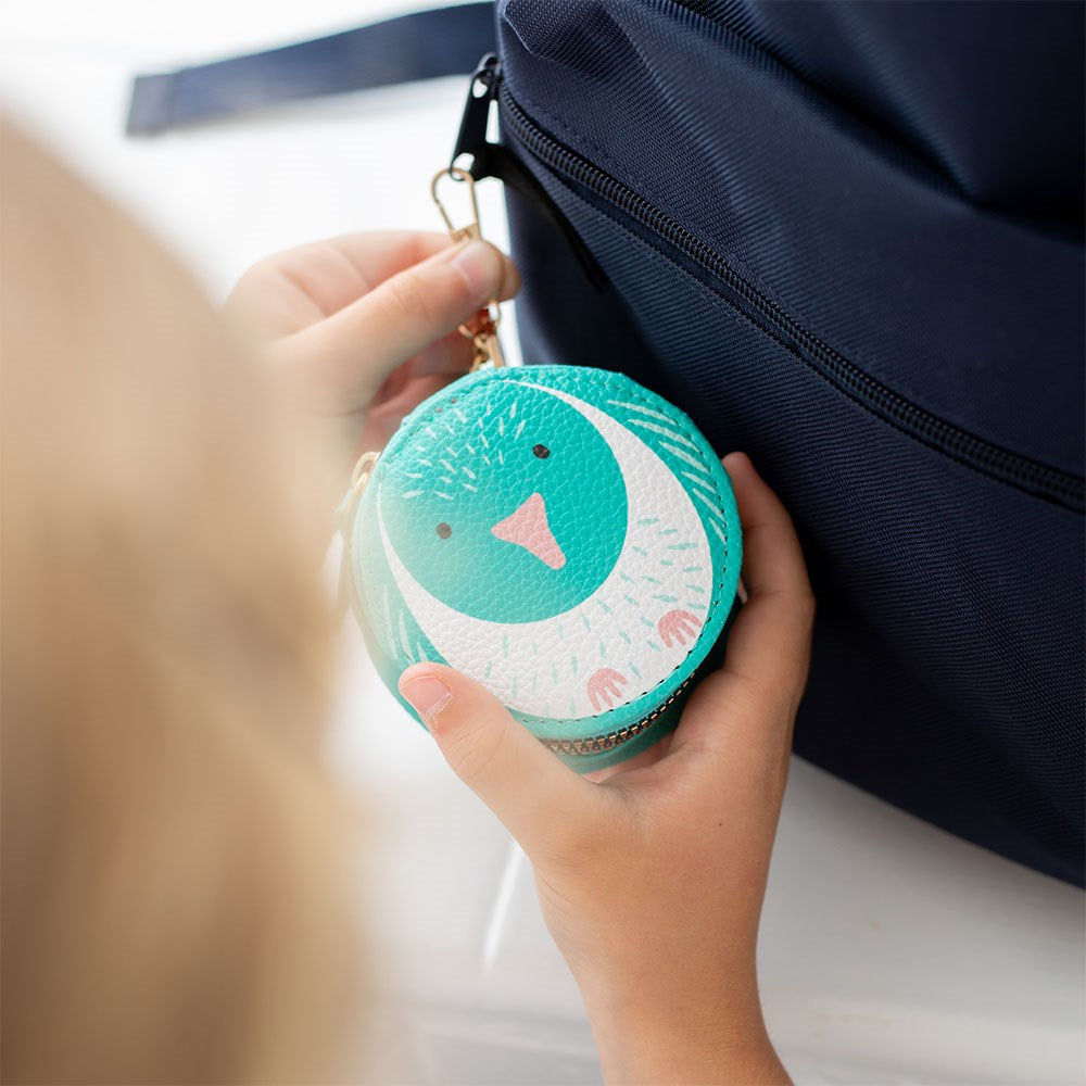 A person holds the Kereru Cutie Coin Purse, a small, round accessory with a teal and white penguin design. It’s attached to a dark blue bag by a gold clasp—perfect for bird lovers.