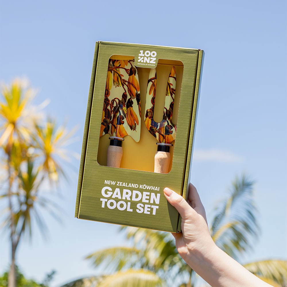 A hand displays the boxed NZ Kowhai Garden Tool Set, featuring floral-patterned tools with ash wood handles. Palm trees and a blue sky appear blurred in the background.