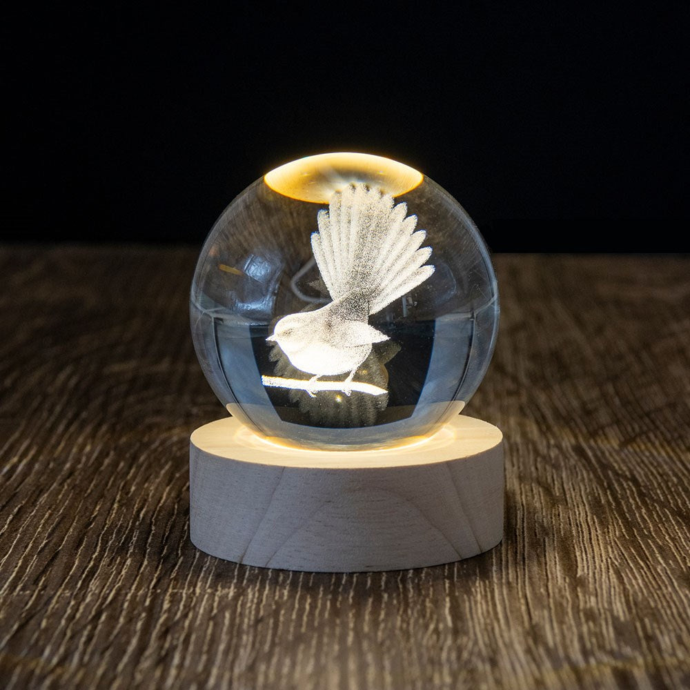 The Fantail LED Globe, a 3D laser-engraved glass sphere with a bird, sits on a lighted wooden base, elegantly displayed on a textured dark surface against a black background.