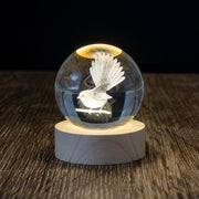 The Fantail LED Globe, a 3D laser-engraved glass sphere with a bird, sits on a lighted wooden base, elegantly displayed on a textured dark surface against a black background.
