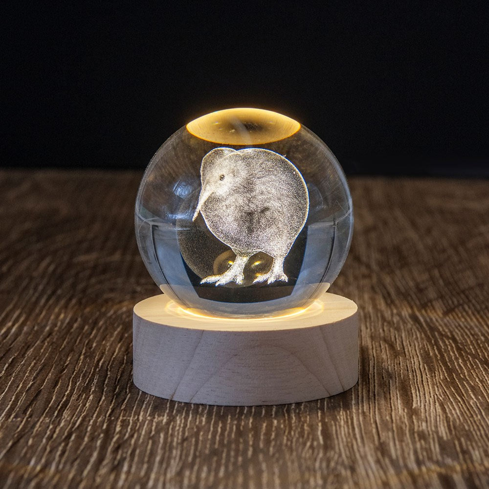 The Kiwi LED Globe is a 3D laser-engraved glass sphere featuring a kiwi bird, set on a round wooden base and illuminated from below. It sits on a textured wooden surface with a dark background, highlighting its glowing design.