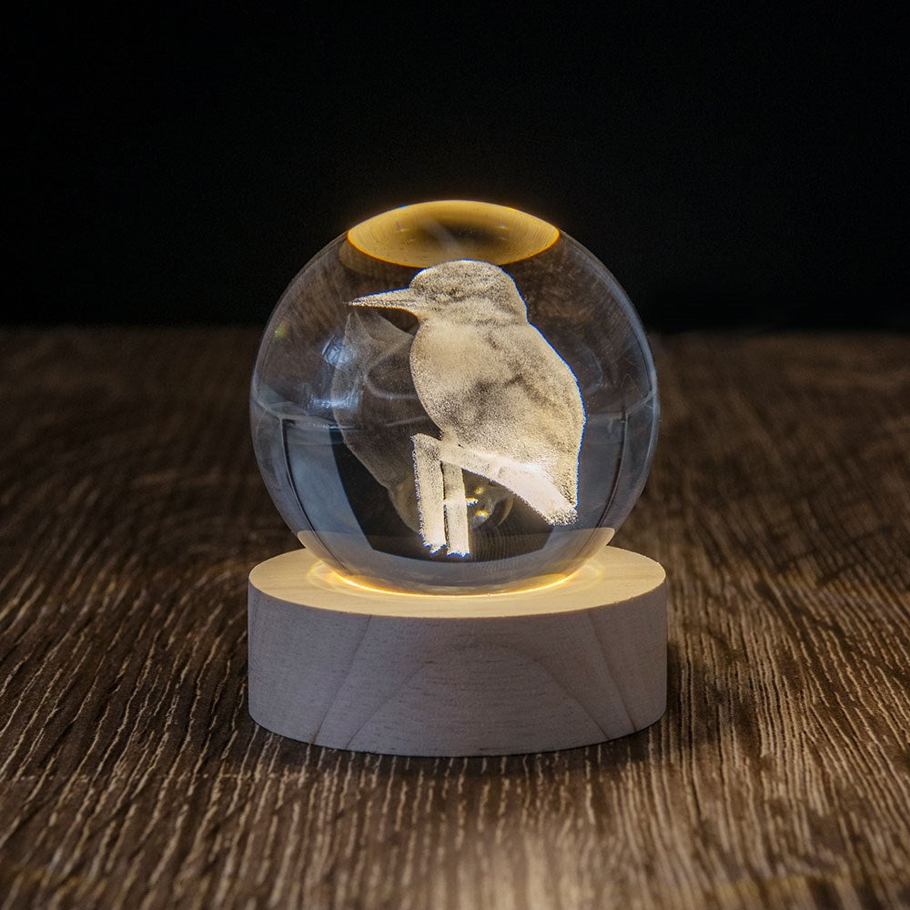 The Kingfisher LED Globe features a glass sphere with a 3D laser-engraved kingfisher, displayed on a round wooden base and illuminated from below, creating a stunning effect against a dark background.