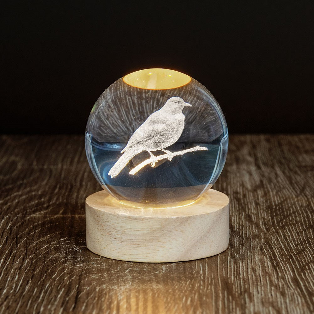 The Tui LED Globe features a glass sphere with a 3D laser-engraved tui on a branch, set on a wooden base. A soft yellow light shines from below, illuminating the bird and branch against darker surroundings.