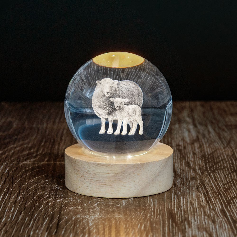 The Sheep LED Globe is a 3D laser-engraved glass sphere with a sheep and lamb, displayed on a wooden LED base atop a dark wood surface against a black background.