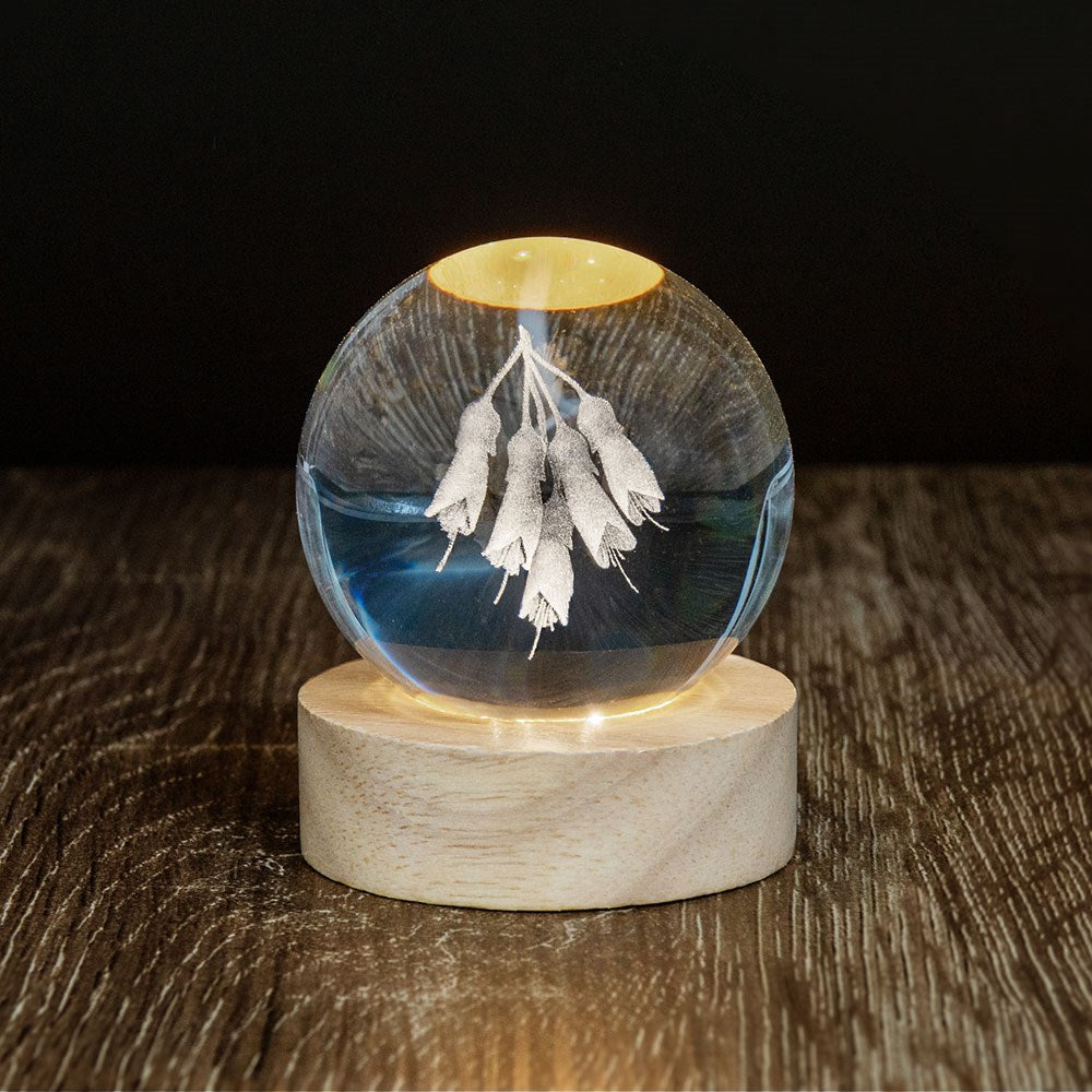 The Kowhai LED Globe features a glass sphere with white, leaf-like designs suspended inside, set on a round wooden base. The 3D laser engraving creates an enchanting display, beautifully highlighting the wood’s grain against a dark background.