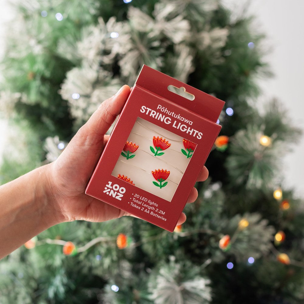 A hand holds a box of Pohutukawa LED Lights—battery powered and designed in New Zealand—in front of a Christmas tree decorated with glowing white fairy lights. The red packaging shows small, red, flower-shaped LEDs.