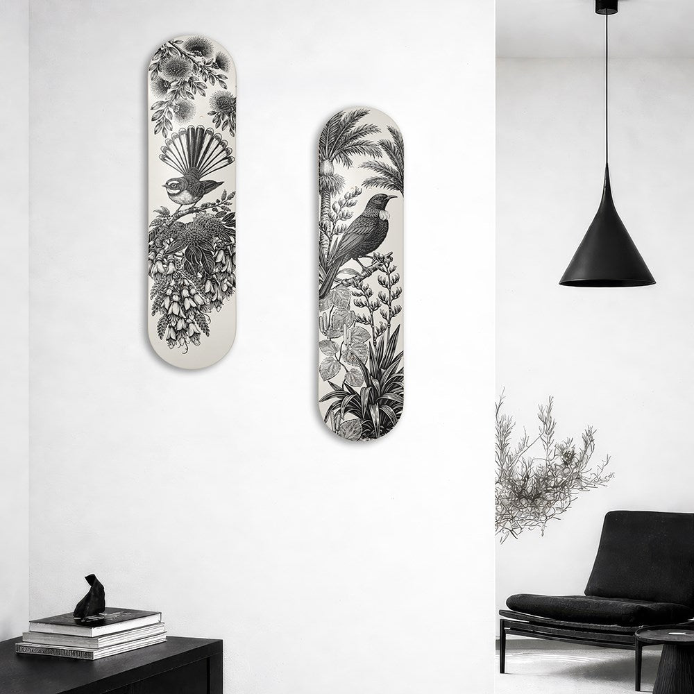 A modern living room with a black lounge chair, black pendant lamp, and two vertical "Under the Nikau Skateboard Deck" wall art pieces featuring detailed bird and NZ flora illustrations. A sculptural object sits on a small table in the foreground.