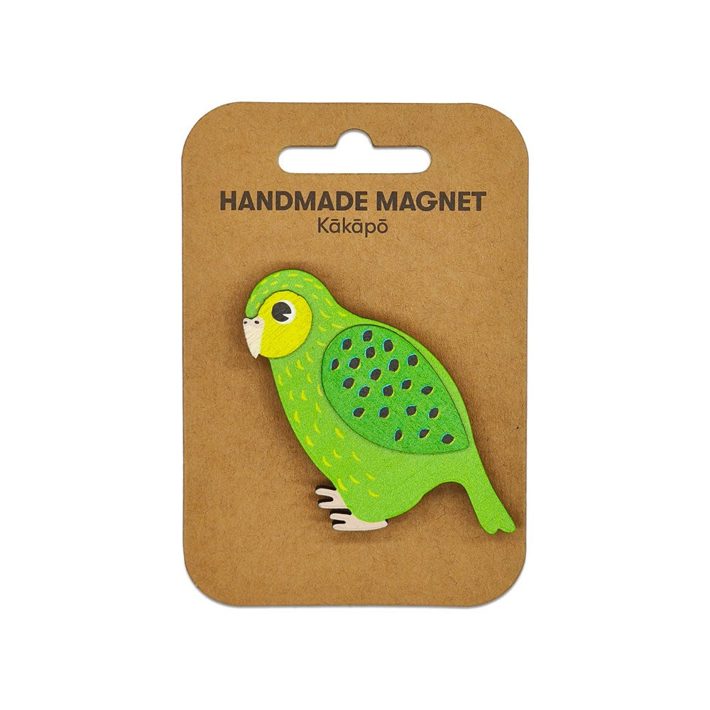 The Kakapo Wooden Magnet is handmade, shaped and painted to look like a green kākāpō bird, and comes attached to a brown card labeled HANDMADE MAGNET Kākāpō with a top cut-out hole for easy hanging.