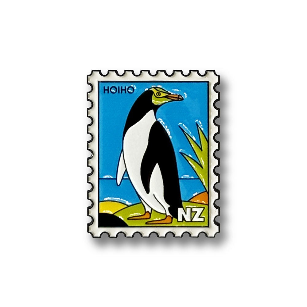 Introducing the Hoiho Collectible Stamp Magnet: a stylized postage stamp illustration featuring a yellow-eyed penguin with "HOIHO" at the top and "NZ" at the bottom right, set against a blue sky and lush greenery. Crafted as an enamel magnet, this charming piece is a perfect collectible and an ideal New Zealand souvenir.