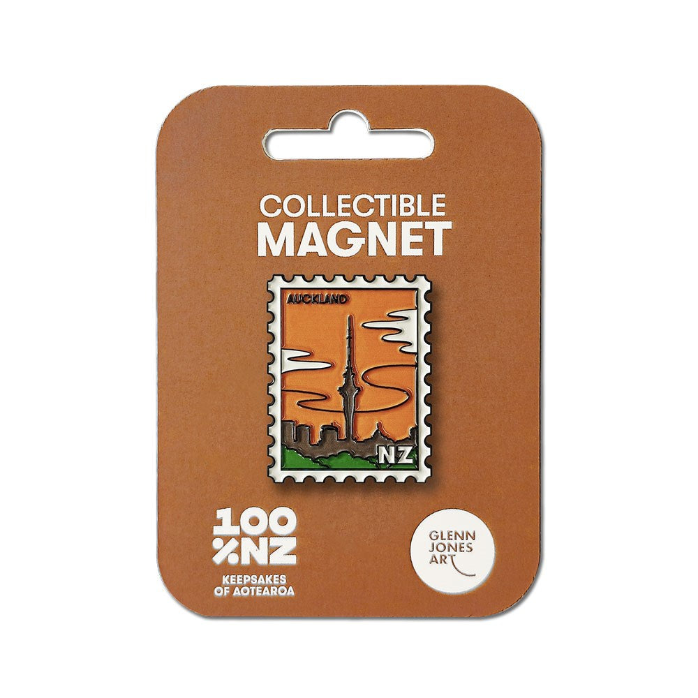 The Auckland Collectible Stamp Magnet showcases Glenn Jones artwork of the citys skyline and Sky Tower on an orange background, shaped like a postage stamp and mounted on a brown card labeled “Collectible Magnet” and “100% NZ.”.