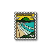 The Mt Maunganui Collectible Stamp Magnet features an illustrated coastal scene with a green mountain, sandy beach, and blue water. Designed by Glenn Jones, it displays NZ in the corner and has a scalloped white border.