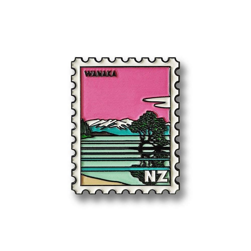 The Wanaka Collectible Stamp Magnet features a landscape with a tree by a lake, mountains, and a pink sky. Designed in New Zealand, it displays “WANAKA” and “NZ.”.