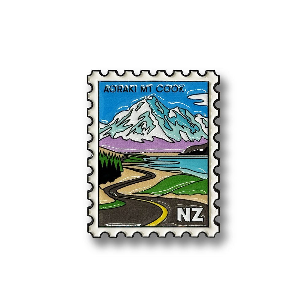 The Aoraki Mt Cook Collectible Stamp Magnet features an illustration of a postage stamp depicting a winding road, lush green trees, a tranquil lake, and the snow-capped peak of Aoraki Mt Cook. “Aoraki Mt Cook” is displayed at the top, with “NZ” in the bottom right corner. Inspired by New Zealand’s breathtaking landscapes, this design is available as a collectible enamel magnet.
