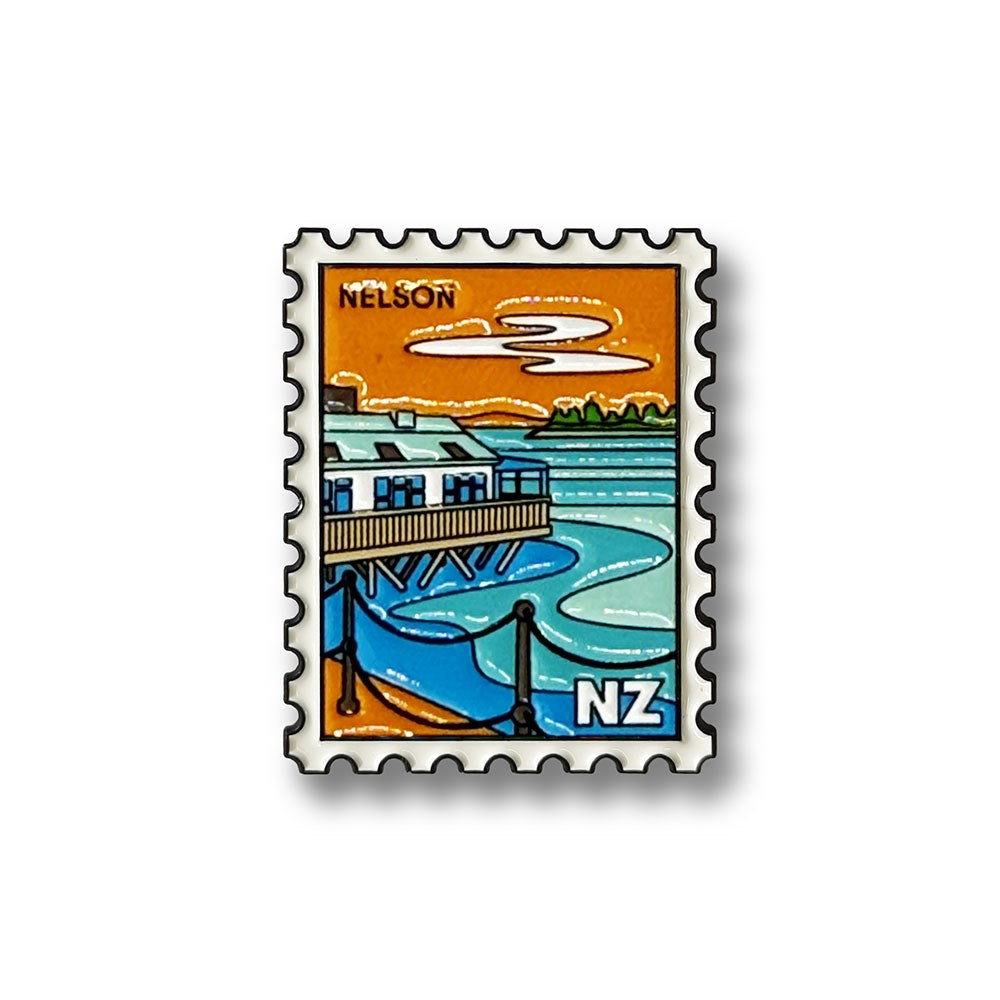 The Nelson Collectible Stamp Magnet showcases a vibrant postage stamp illustration of a waterside building with a pier, blue water, and an orange sky. Featuring "NELSON" at the top and "NZ" at the bottom right to represent Nelson, New Zealand, this unique enamel magnet is a perfect collectible for anyone who enjoys distinctive New Zealand keepsakes.