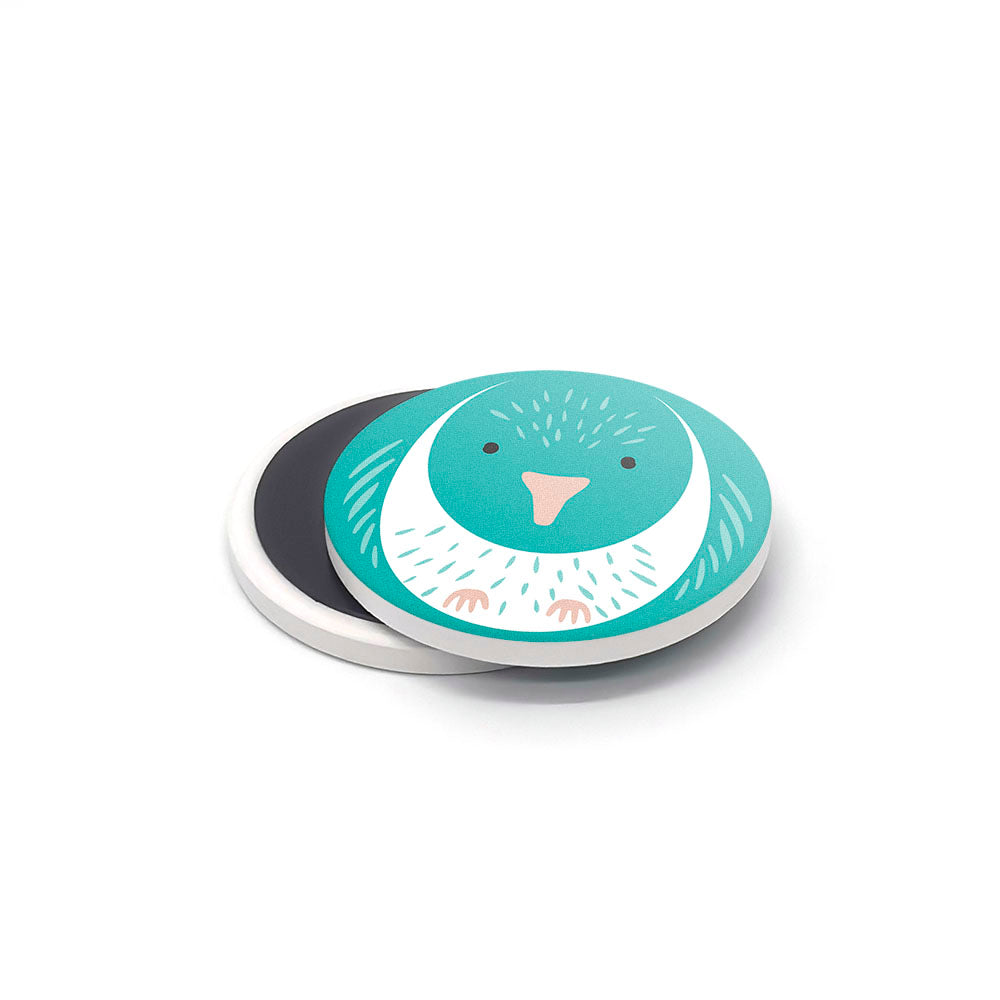 The NZ Cuties Ceramic Magnet Set includes a round magnet with a teal and white bird illustration, shown on a white surface partly overlapping a black magnet—ideal for anyone searching for distinctive New Zealand souvenirs.